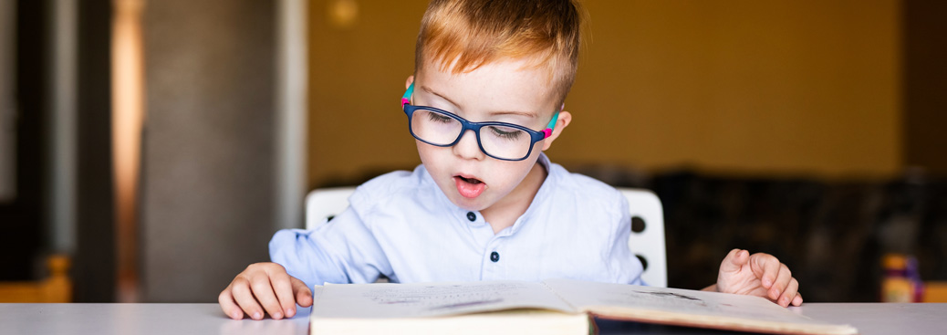 Image of a young male primary school student wearing glasses sitting at a table reading a book